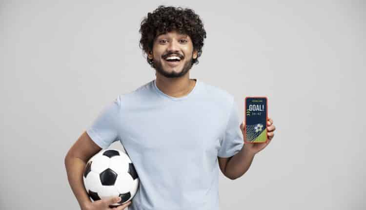 Homem segurando uma bola em um dos braços e, na outra mão, mostrando um celular.